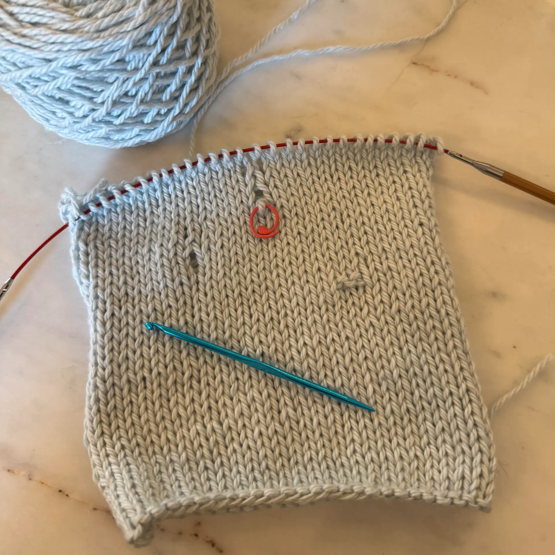 Light-colored knitted swatch on circular needles with a stitch marker and crochet hook